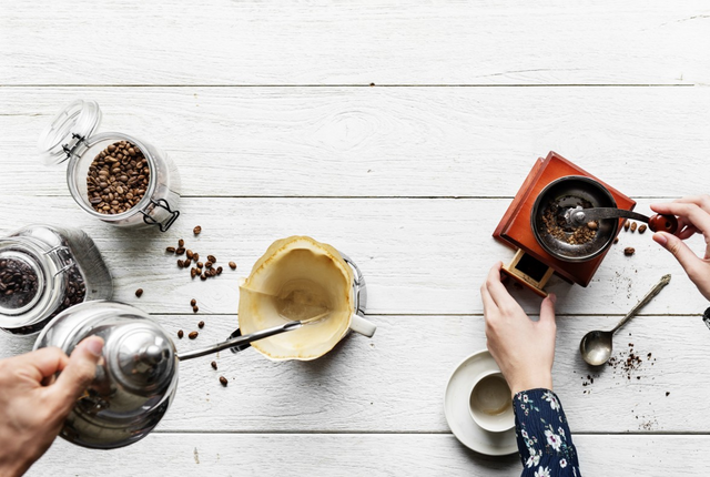 Mehrere Händen umfassen über einem Tisch eine Kaffeekanne und eine Kaffeemühle. Drumherum stehen Kaffeeutensilien wie Tassen und Bohnen.