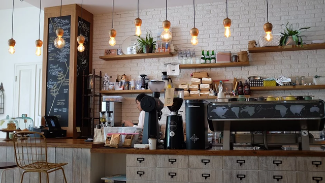 Eine Kaffeebar mit hölzerner Theke, einem Kaffeevollautomaten und einem beschäftigten Barista dahinter. Das Café wirkt hell und freundlich, Lampen hängen von der Decke und es gibt eine große Auswahl an Speisen und Getränken.