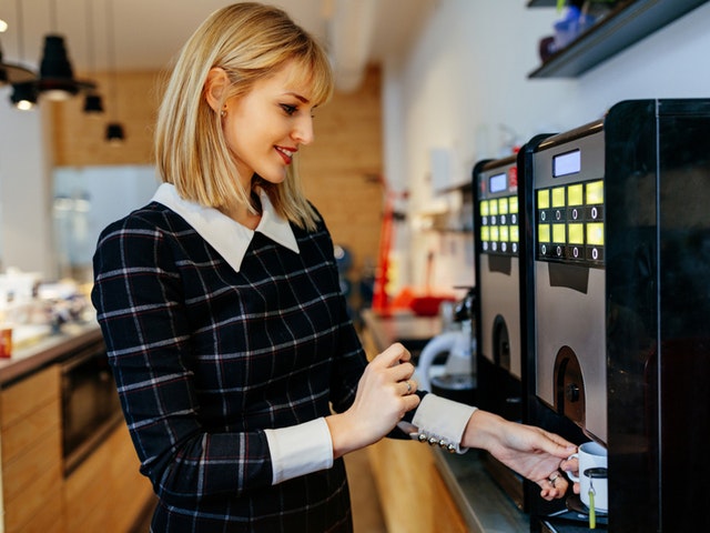 Eine junge Frau in Bürokleidung holt sich einen Kaffee am Vollautomaten